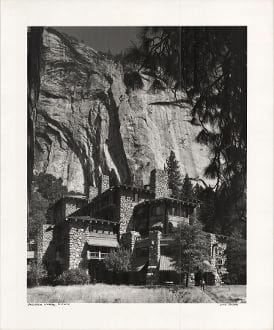 A black and white photo of a house in front of a rock face.