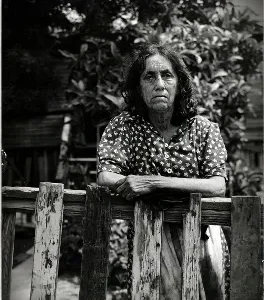 An elderly woman stands behind a wooden fence in a garden.